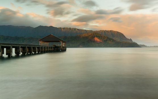 Rising sun illuminates the peaks of Na Pali mountains over the calm bay and Hanalei Pier in long exposure photo