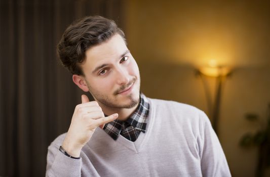 Attractive young man doing "call me" sign with hand and fingers, cheerful expression looking at camera