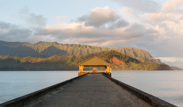 Rising sun illuminates the peaks of Na Pali mountains over the calm bay and Hanalei Pier in long exposure photo