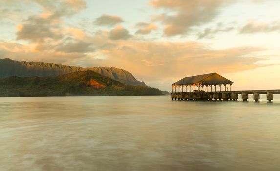Rising sun illuminates the peaks of Na Pali mountains over the calm bay and Hanalei Pier in long exposure photo