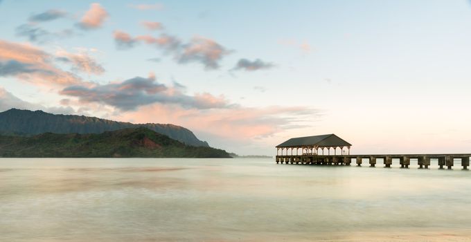 Rising sun illuminates the peaks of Na Pali mountains over the calm bay and Hanalei Pier in long exposure photo