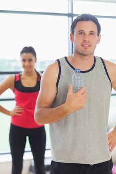 Portrait of a fit young man holding water bottle with friend in background in a bright exercise room