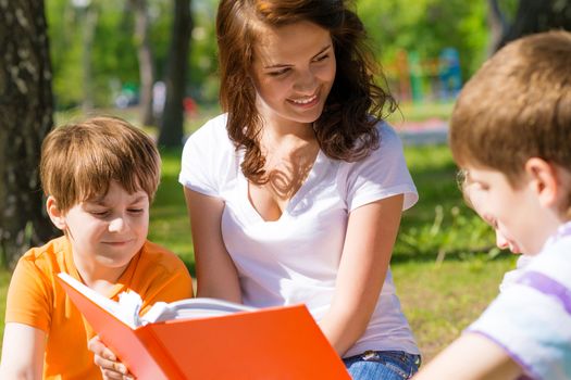 children and teacher reading book together in the summer park