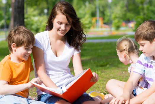 children and teacher reading book together in the summer park