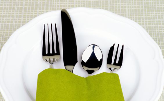 Elegant Table Setting with Fork, Table Knife, Spoon and Dessert Fork into Green Napkin Decorated on White Plate closeup on Checkered background 