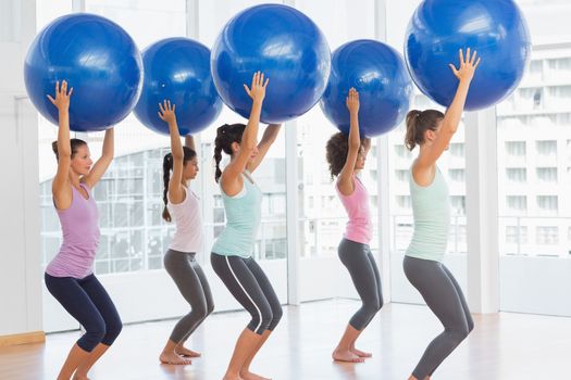 Full length side view of fit young women holding blue fitness balls in exercise room