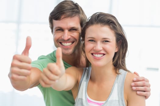 Portrait of a fit young couple gesturing thumbs up in bright exercise room