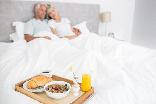 Blurred mature couple sitting on bed with breakfast in foreground at home