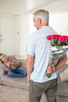 Rear view of a man holding bouquet behind his back with woman sitting on couch at home