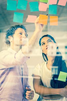 Businessman and businesswoman discussing over adhesive notes on glass wall in office