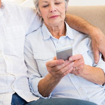 Senior woman reading text message on smartphone while sitting with man at home