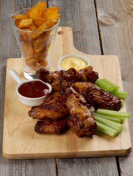 Juicy Chicken Legs and Wings Barbecue with Ketchup, Cheese Sauce, Celery Sticks  and French Fries in Glass closeup on Wooden Cutting Board