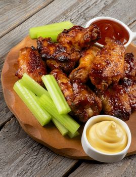 Spicy Chicken Legs and Wings Barbecue with Celery Sticks, Ketchup and Cheese Sauce on Wooden Plate closeup on Rustic Wooden background