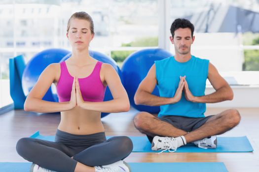 Full length of a young couple sitting with joined hands and eyes closed at fitness studio