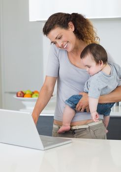 Smiling mother using laptop while carrying baby boy at kitchen counter