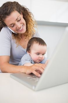 Happy woman using smartphone and laptop while sitting with baby boy at counter