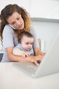 Mother using laptop and cellphone while sitting with baby at counter 