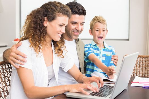 Laughing little boy using laptop with parents at table at home in kitchen