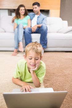 Cute little boy using laptop on the rug with parents sitting sofa at home in living room