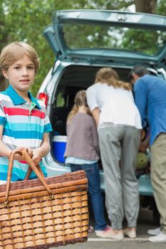 Portrait of a boy with picnic basket while family in background at car trunk