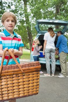 Portrait of a boy with picnic basket while family in background at car trunk