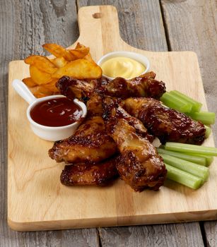Juicy Chicken Legs and Wings Barbecue with Ketchup, Cheese Sauce, Celery Sticks  and Heap of French Fries closeup on Wooden Cutting Board