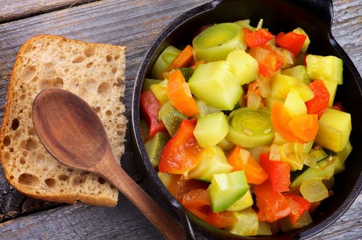 Vegetable Stew in Black Saucepan with Brown Bread and Wooden Spoon closeup on Rustic background. Top View