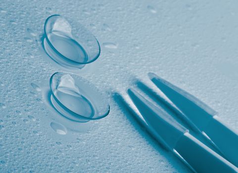 Arrangement of Two Contact Lenses with Water Droplets and Special Tweezers isolated on Wet background. Blue Toned