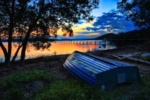 Beautiful sunset landscape with  blue weathered upturned boat at shoreline at low tide.  This pretty spot is Kincumber, Australia.  Please note long exposure, there is some motion in trees due to slight breeze