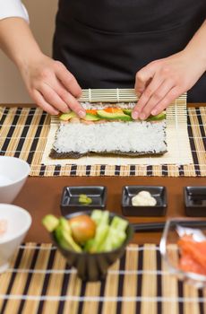 Hands of woman chef rolling up a japanese sushi with rice, avocado and shrimps on nori seaweed sheet