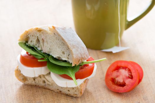 Healthy nutritious breakfast consisting of a sandwich made of potato bread, mozzarella cheese, tomatoes and fresh leaves of baby spinach, next to a cup of tea, on a wooden table