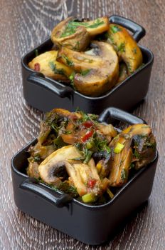Delicious Roasted Edible Champignon Mushrooms with Onion and Greens in Black Cast-iron Stew Pots isolated on Wooden background