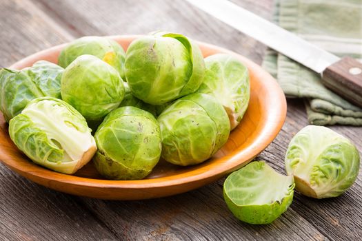 Bowl of fresh uncooked cleaned brussels sprouts with one halved one in the foreground standing on an old rustic wooden kitchen table alongside a knife