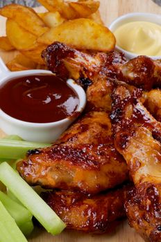 Juicy Chicken Legs and Wings Barbecue with French Fries, Ketchup, Cheese Sauce and Celery Sticks closeup on Wooden Board