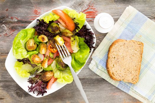 Delicious fresh leafy green mixed salad with diced and sauteed brussels sprouts served on a rustic wooden table with a slice of brown bread for a healthy lunch, overhead view
