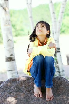 Young biracial girl quietly sitting on rock under trees outdoors in summer, peaceful scene