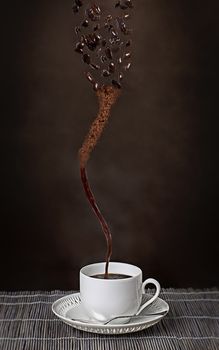 Cup of espresso with a whirlwind of coffee beans becoming a stream of ground coffee powder before finally evolving into a cup of hot freshly brewed full roast espresso in a conceptual image