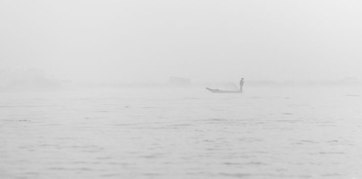 Foggy morning weather on a lake with silhouette of fisherman in small wooden boat 