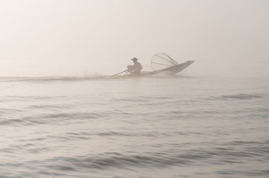 Foggy morning weather on a lake with silhouette of fisherman in small wooden boat with motor