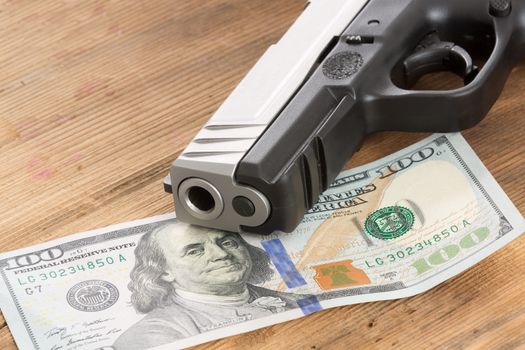 Close up of the muzzle of a gun with a 100 American dollar bill lying on a wooden table in a conceptual image of crime, bribery and corruption