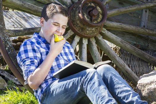 Relax.Young boy reading a book in the woods eating an apple
