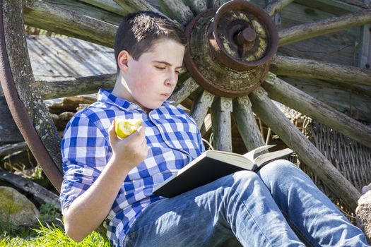 Relax.Young boy reading a book in the woods eating an apple