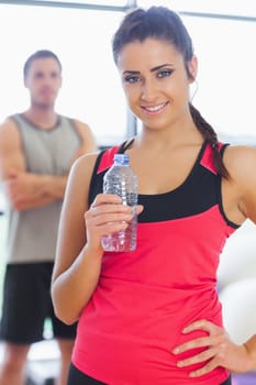 Portrait of a fit young woman holding water bottle with friend in background in a bright exercise room