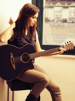 Photo of a woman playing an acoustic guitar while sitting by a window.