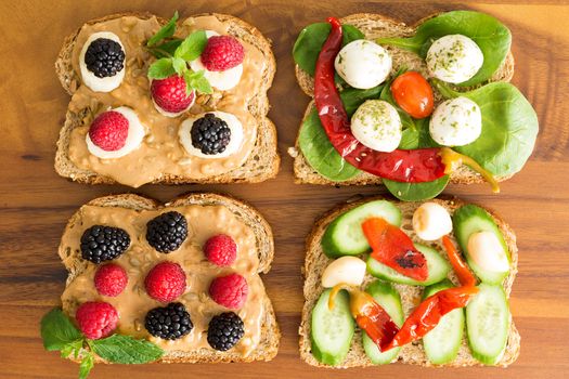 Four healthy open sandwiches for a picnic lunch topped with fresh berries, cheese, chili peppers, cucumber and baby spinach on wholewheat bread, overhead view on a wooden table