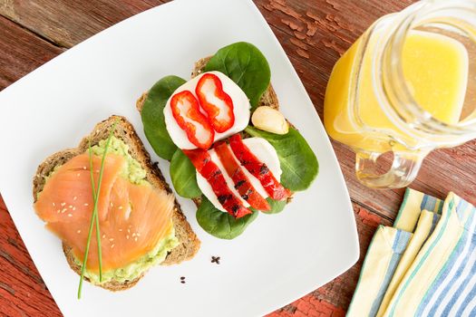 Healthy gourmet snacks for a picnic served on a white plate with wholewheat bread topped with smoked salmon, avocado, mozzarella cheese, sweet peppers and baby spinach and a jug of fresh orange juice