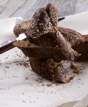 Heap of Sliced Sesame and Poppy Seed Brown Bread with Table Knife on Parchment Paper closeup on Wooden background 