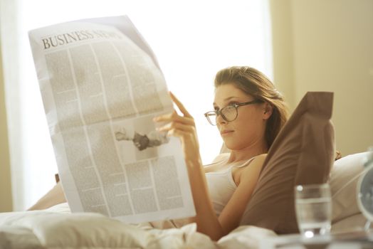 Young woman lying on the bed at home and reading the newspaper 