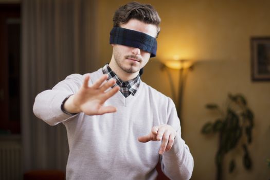 Blindfolded young man at home in living room cannot see, trying to find his way with his hands