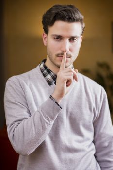 Handsome young man doing Hush sign with finger on lips, looking at camera. On location